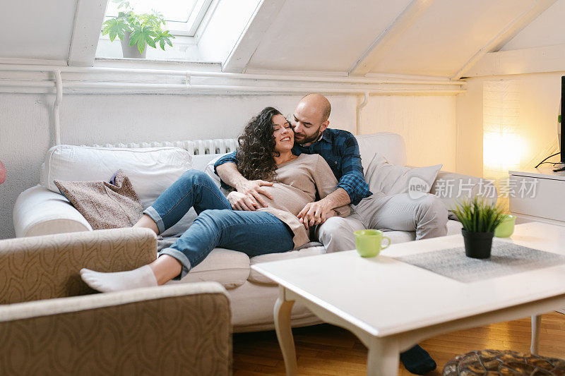 浪漫的新婚夫妇幸福地拥抱在沙发上，同时抚摸着母亲怀孕的肚子