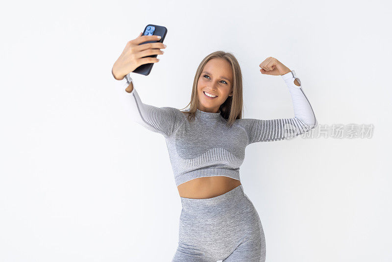快乐的运动女人用智能手机自拍，在白色背景下展示二头肌