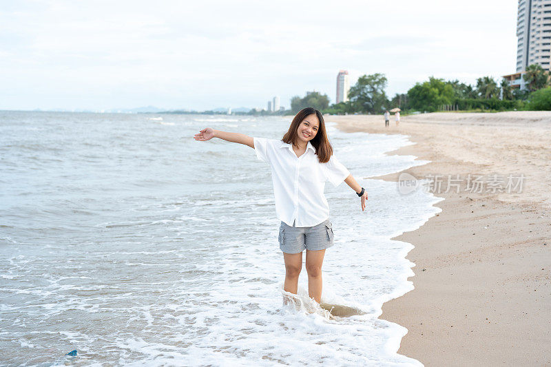 身穿白衬衫、长发的亚洲女性在周末度假时，正带着愉快的表情走在海浪不断拍打的海滩上。日落时的傍晚气氛