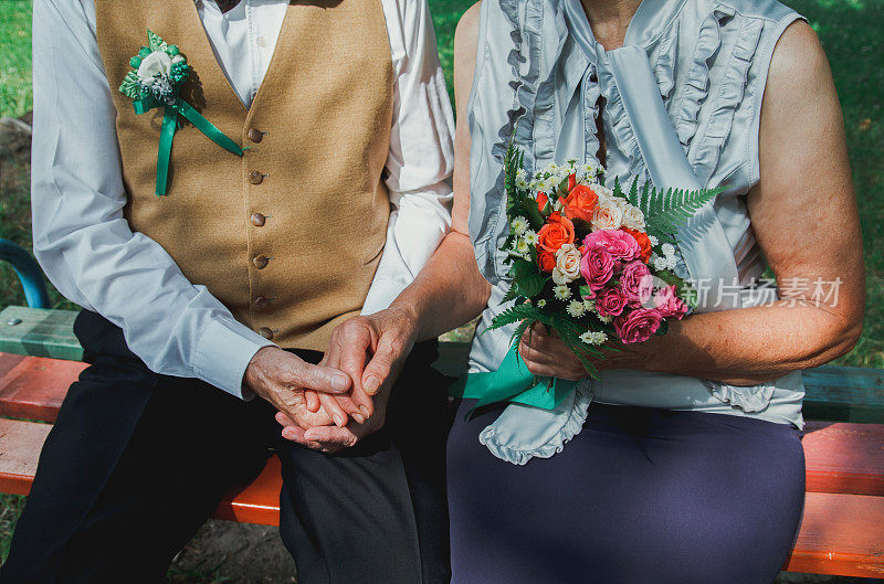 一对老夫妇坐在绿色公园的长凳上。祖母和祖父在他们的金婚纪念日上。五十年在一起。老人牵着手捧着质朴的花束。