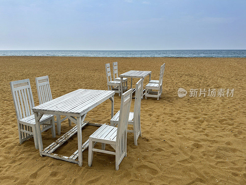 图片中，金色沙滩上质朴的木条餐桌，一组白色的木制桌椅，斯里兰卡科伦坡拉维尼亚山海滩上的游客露天用餐区，蓝色的天空和大海背景，复制空间