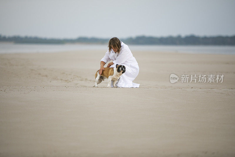 年轻的成年女子和她的法国斗牛犬在海滩上散步