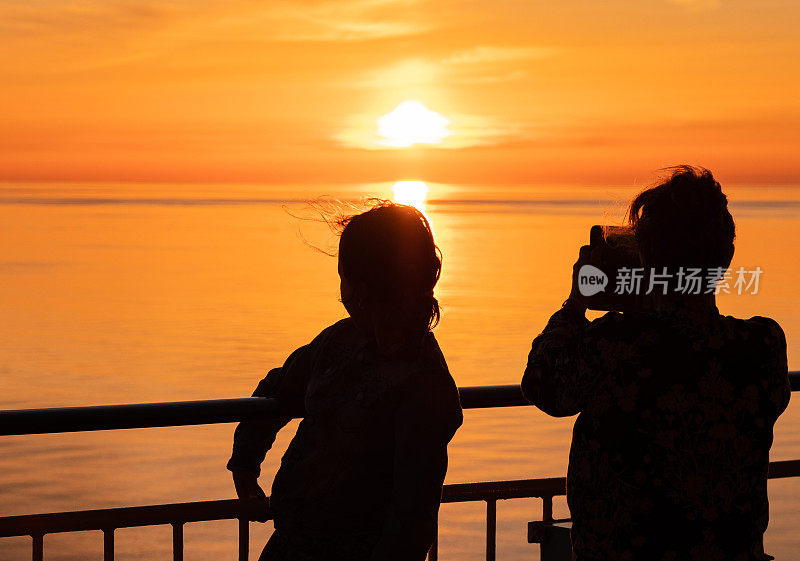 一家人，一个年轻的女人和一个孩子站在船的甲板上，在夕阳的映衬下拍照。小女孩和女人的剪影。