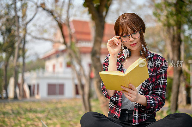 美丽的亚洲女大学生在公园里看书时正在摸眼镜
