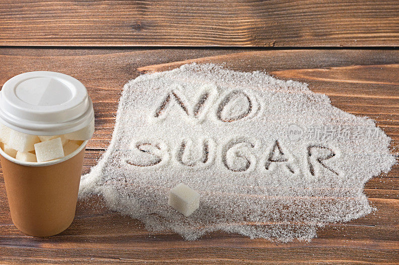 在棕色的背景上，有一个装满精制糖的纸玻璃杯，一堆糖上写着“不加糖”的字样。减少摄入和放弃糖的概念