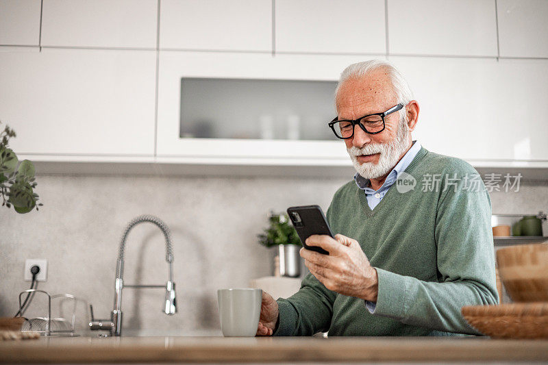 老人，退休老人，坐在厨房里，带着快乐的微笑，喝着热咖啡或茶，用着手机，早晨的例行公事，无忧无虑的退休日子