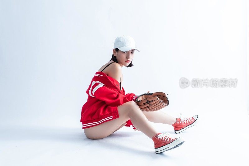 一位戴着棒球帽、穿着夹克和运动鞋的年轻女子坐在地上，手里拿着棒球手套。