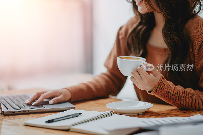 一个女商人的手拿着咖啡杯，在笔记本电脑上打字的特写，表明一天的开始是高效的。