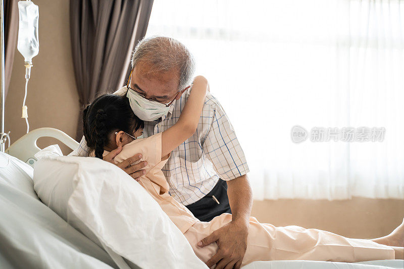 亚洲老爷爷探望并拥抱躺在医院病床上的小女孩。他们戴着口罩，防止来自医院的人感染冠状病毒。