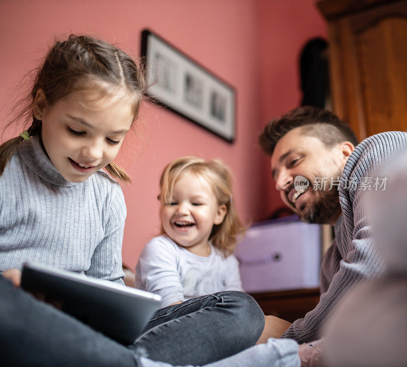 单身年轻父亲躺在床上，和他的女儿们在家里用平板电脑看书