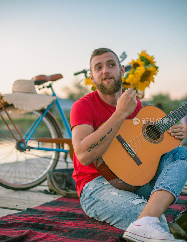 时尚的潮人坐在木码头上，拿着原声吉他对着美丽的湖