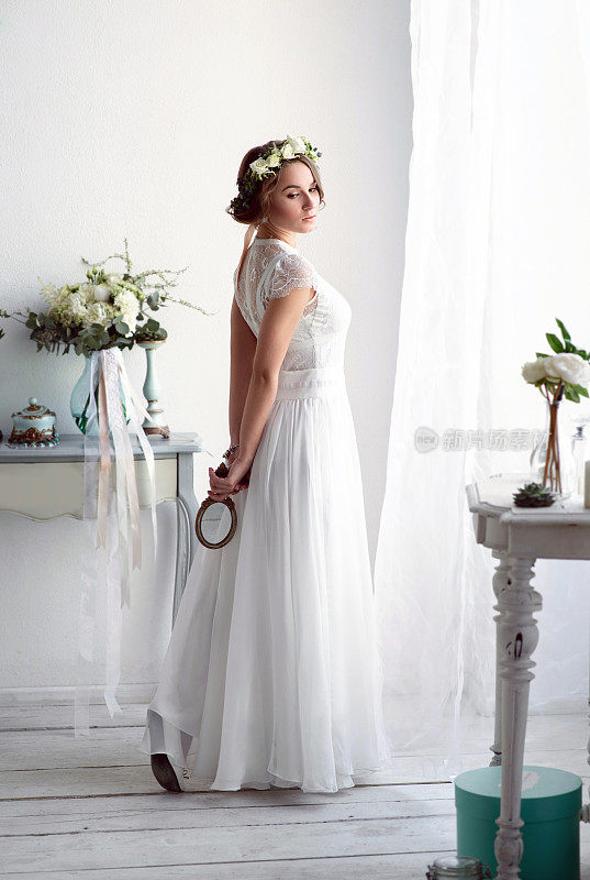 在婚礼当天，新娘穿着优雅的白色礼服靠近窗户。浪漫的早晨，美丽的装饰