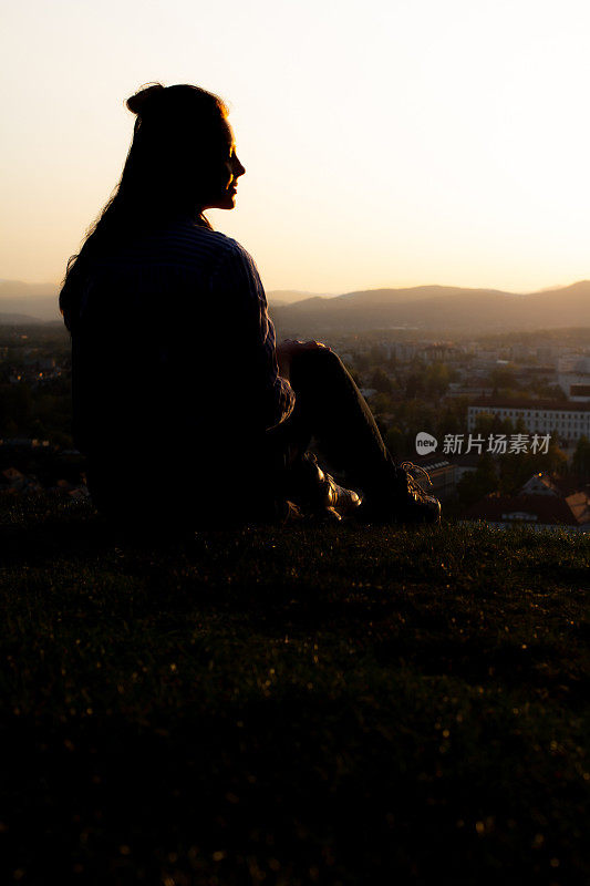 一个女人的黑色剪影坐在草山上俯瞰风景。