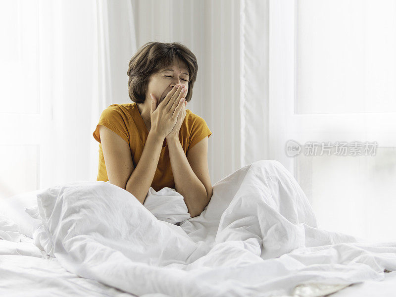 瞌睡的女人盖着白毯子，躺在床上打哈欠。早上很难早起。
