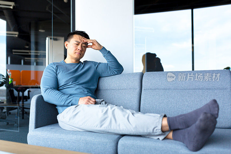 在一间现代化的办公室里，一名疲惫而沉思的亚洲工人躺在办公室的沙发上，一位商人做出了一个艰难而致命的决定
