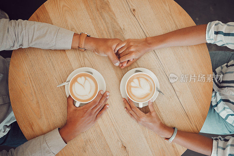 在咖啡馆里，一对相爱的年轻情侣手牵手，桌上放着两杯咖啡。男性和女性在约会时从上面用热卡布奇诺或浓缩咖啡手
