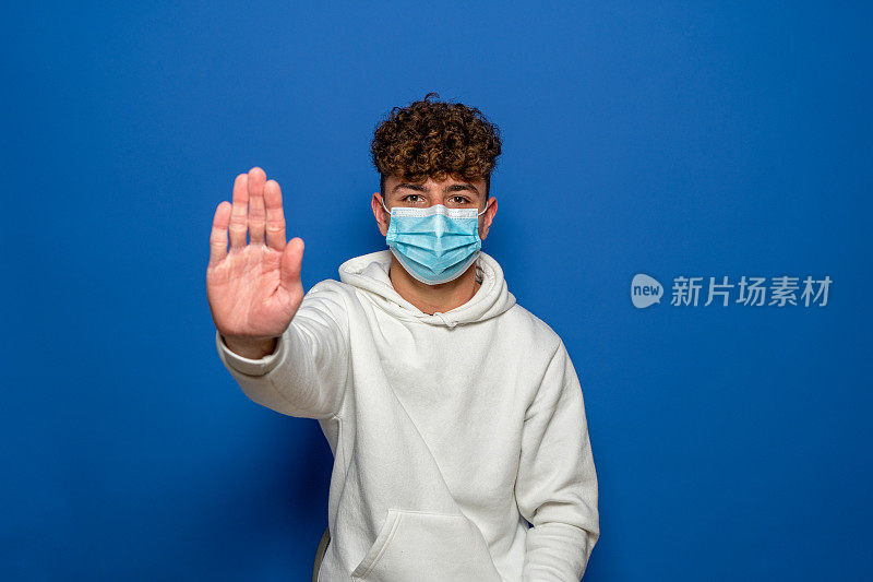 白人青少年戴着面具在蓝色工作室背景上做停止手势。青少年对冠状病毒大流行持否定态度，不满新常态的生活方式。