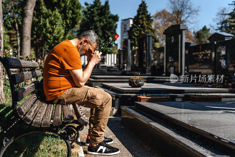 成熟悲伤的男人坐在墓地的墓碑旁，手里拿着一朵花，哀悼死者