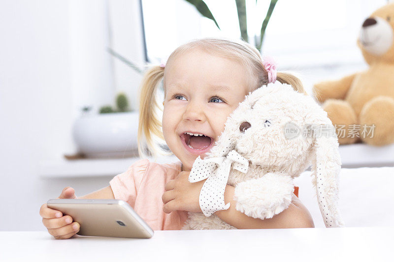 女孩带着玩具兔用手机，用智能手机打视频电话，和亲戚聊天，女孩坐在家里，用电脑在线摄像头，打视频电话。