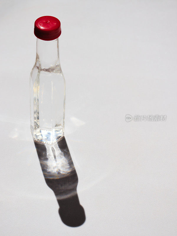红色瓶盖的透明玻璃瓶，里面装着无色液体，晨光从侧面照亮，白色表面投射出阴影和倒影。
