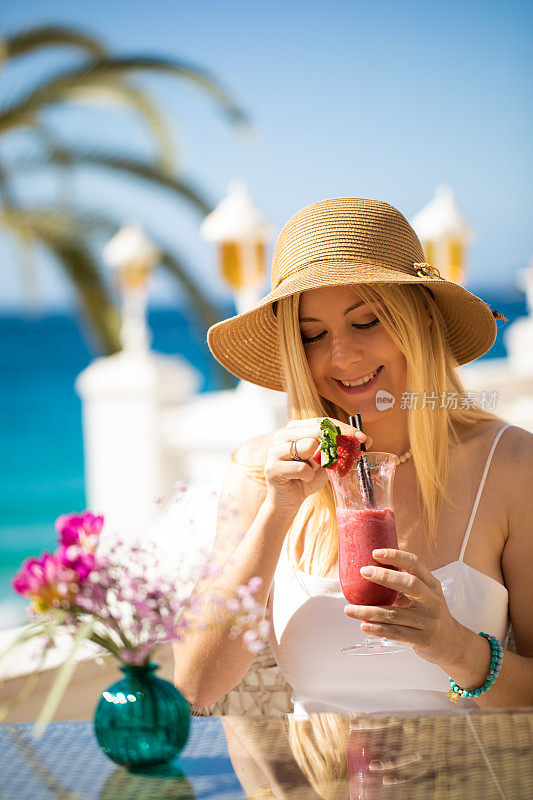 年轻的金发女郎坐在海滩餐厅的桌子上，享受着蓝绿色大海的红色鸡尾酒