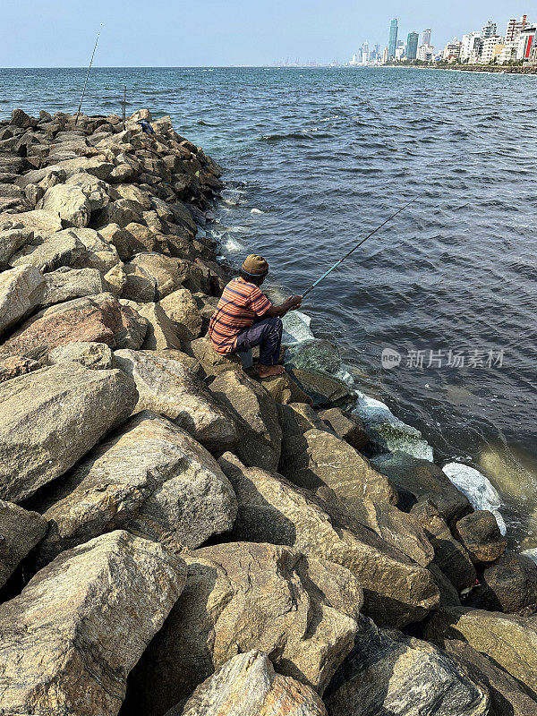 渔夫坐在海岸线上的岩石自然海防上的鱼竿特写图像，海岸侵蚀抛石岩石装甲墙，海岸管理，岩石堆，海滩岩石装甲，海浪，背景中的城市摩天大楼，科伦坡，斯里兰卡