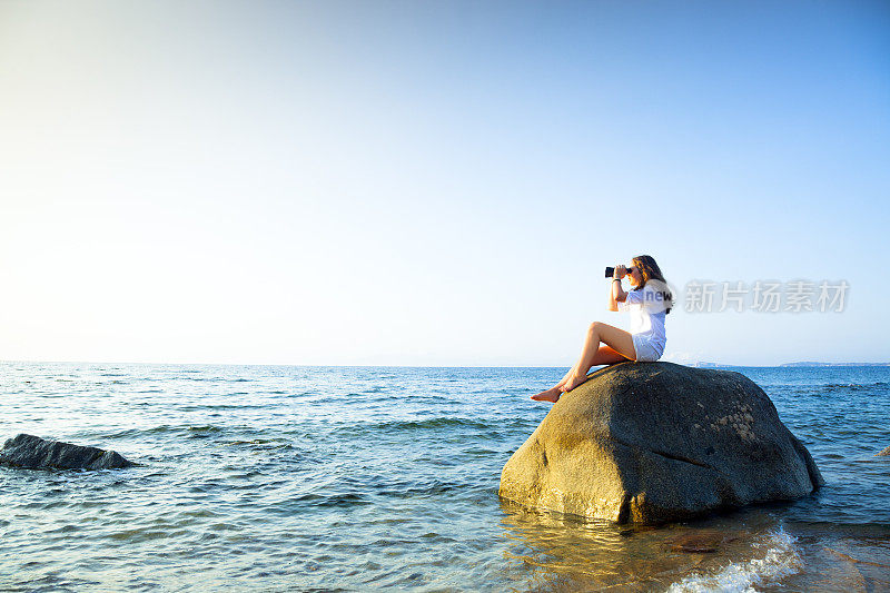 一个少年拿着双筒望远镜坐在海里的一块岩石上。