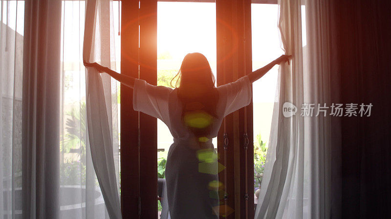 穿着浴袍的年轻女子拉开窗帘，伸懒腰，站在家里靠近窗户的地方。