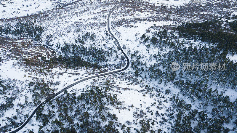 在恶劣天气下，俯视图无人机在弯曲的道路、风山和覆盖着积雪的森林上空飞行。