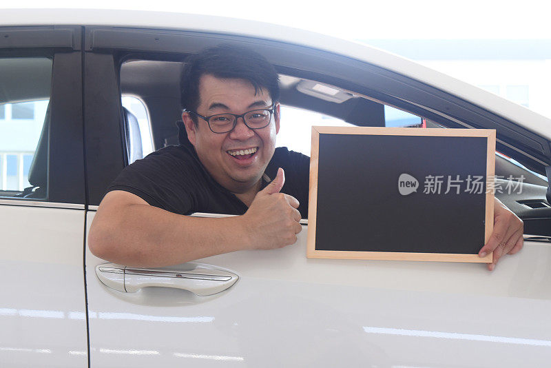 一名亚裔男子坐在车里，手里拿着一张黑色空白海报，竖起大拇指。