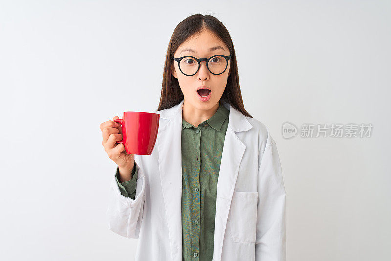 年轻的中国女医生戴着眼镜，在孤立的白色背景下喝咖啡，在震惊中吃惊的脸，害怕和兴奋与恐惧的表情
