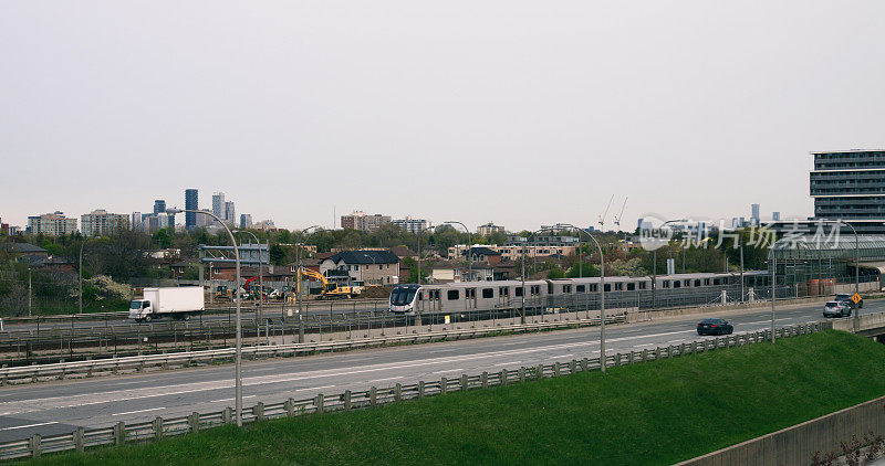 地铁列车从城市沿着住宅区的铁路行驶。广阔的城市景观与火车，高速公路与飞驰的汽车，房屋，绿色草地。概念使用火车准时到达