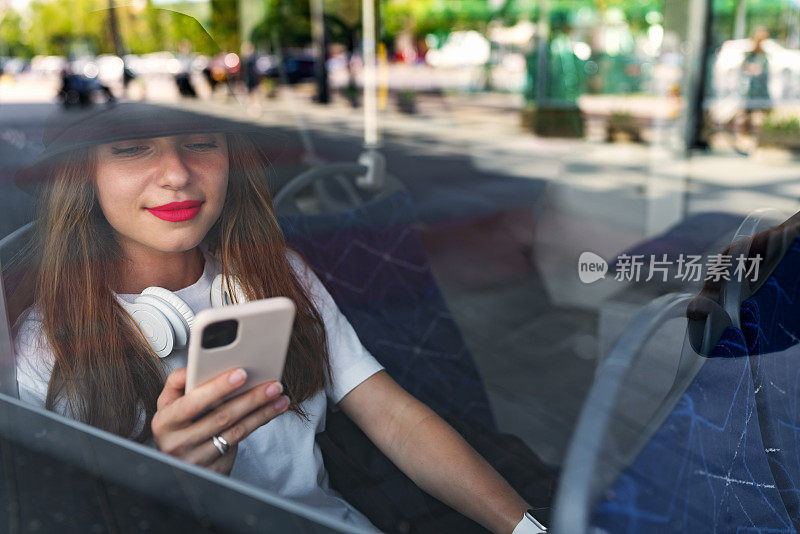 透过巴士玻璃窗拍摄的照片，女乘客坐在穿梭巴士上使用智能手机。