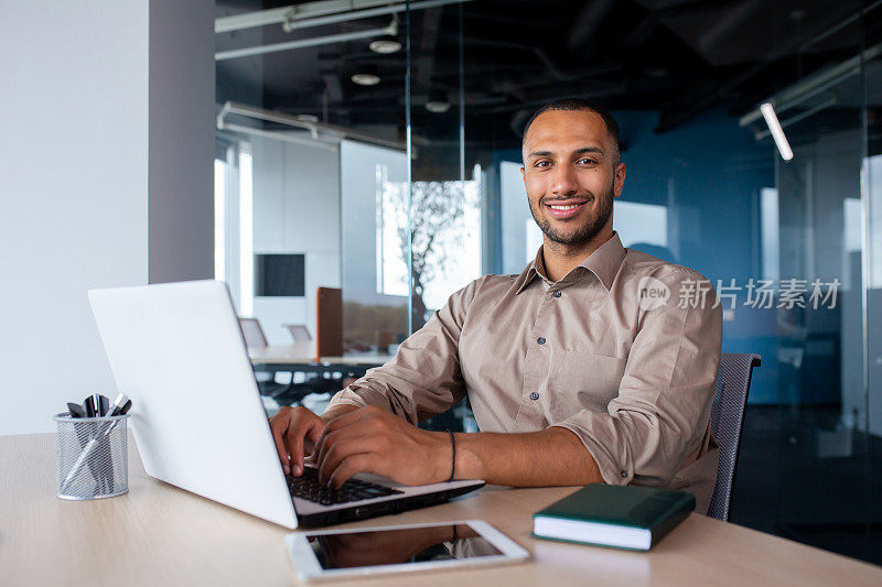 有魅力的非洲裔美国人坐在办公桌前的笔记本电脑在办公室，程序员在现代环境下工作，敲击键盘和微笑的镜头