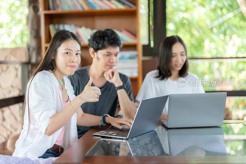 亚洲女学生和两个朋友在笔记本电脑上在线学习的侧视图，竖起大拇指，像大学图书馆的标志一样，每周集体读书以获得知识和智慧。