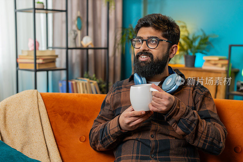 迷人的微笑印度年轻人早上在家里的沙发上喝一杯咖啡或花草茶