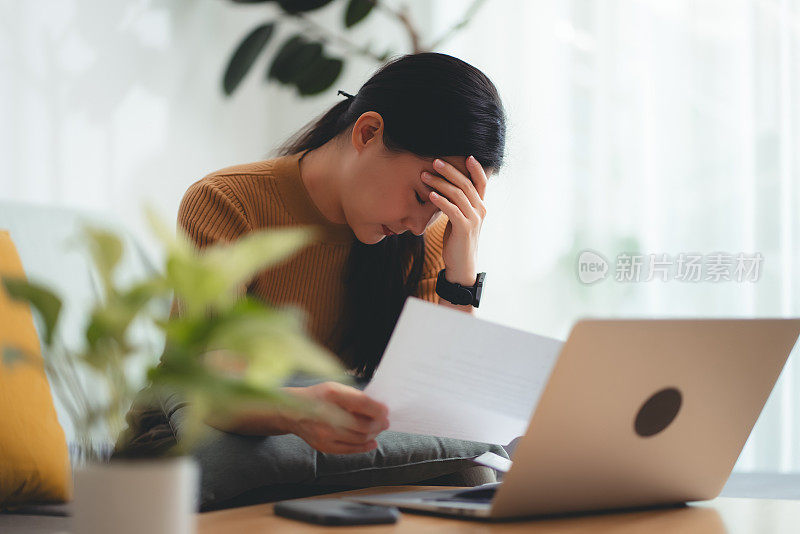 亚洲女性坐在沙发上，担心银行票据、抵押贷款债务和生活成本的账单支付问题。