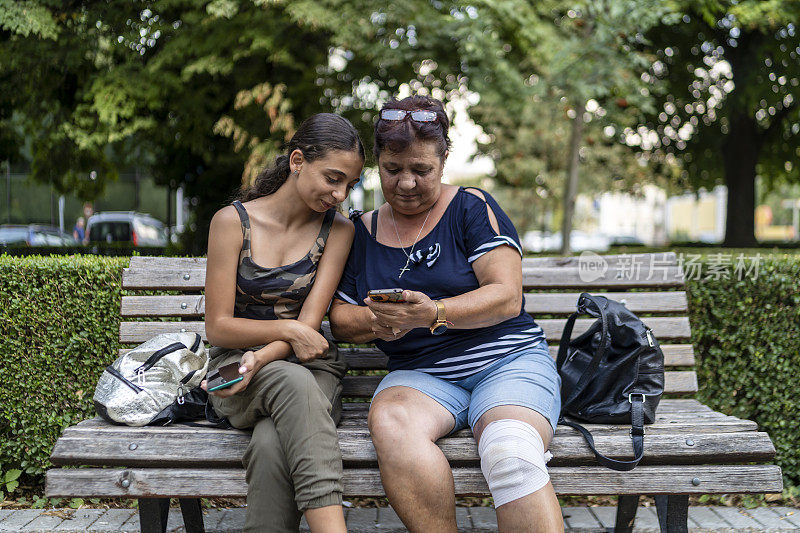 一个十几岁的女孩在公园的长椅上向她的祖母展示如何使用电话