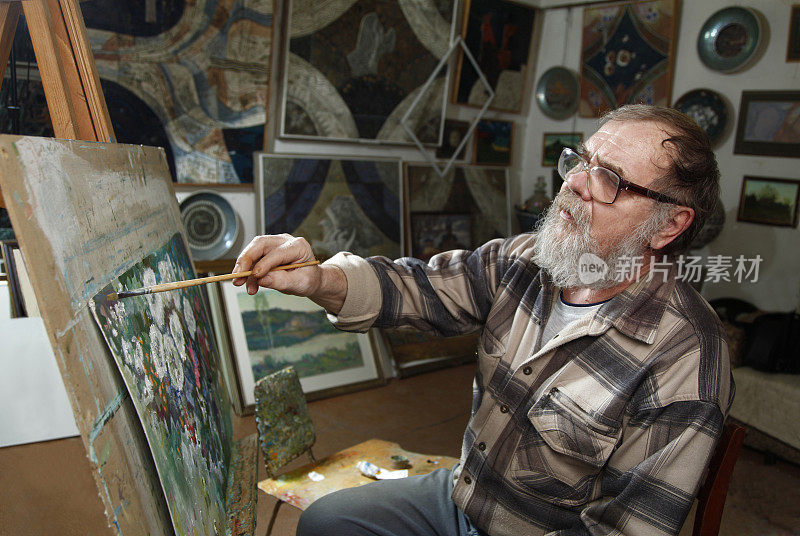 一位戴着眼镜，留着胡子的老画家在艺术工作室用油画画一幅花
