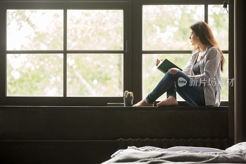 沉思的梦幻女孩拿着书坐在家里的窗台上