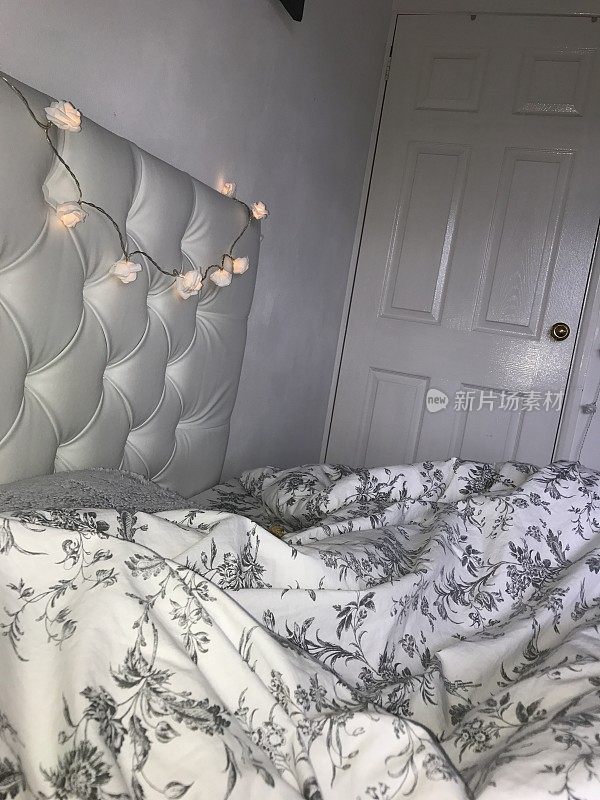 家庭卧室里的双人床，白色镶钉的软垫床头板装饰着仙女灯，皱褶的羽绒被和灰色的散垫，白色和灰色的室内卧室设计照片