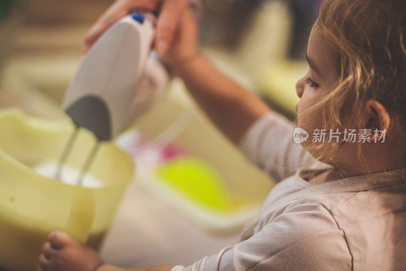 一个蹒跚学步的孩子协助她的母亲在搅拌一个蛋糕面糊与电动搅拌机