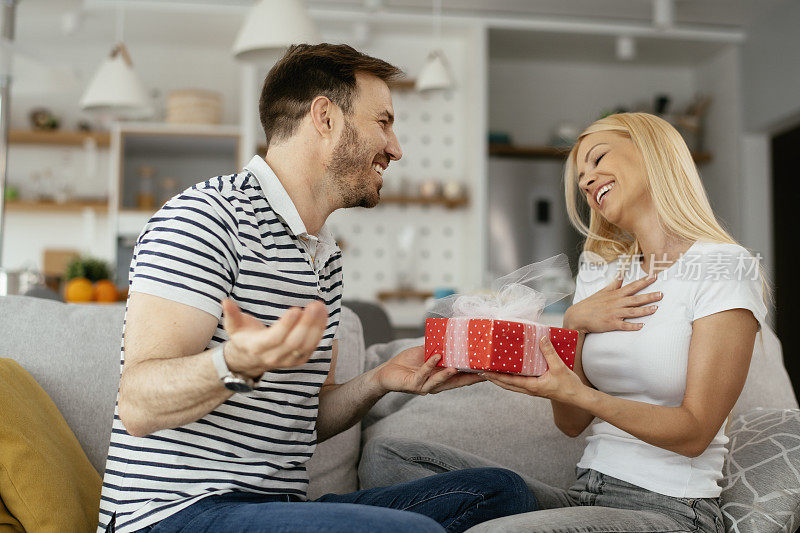 微笑的男人惊喜他的女朋友在家里的礼物。库存图片