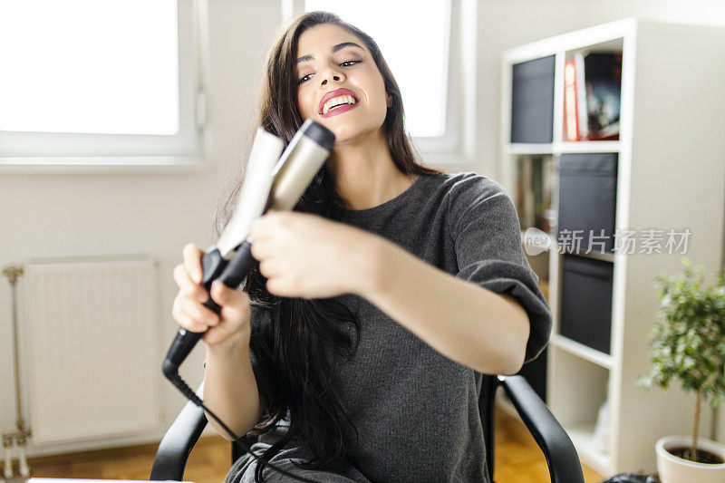 坐在梳妆台边看电脑边用卷发棒的女人。在冠状病毒大流行的时候，一名妇女通过在网上看教程来做头发