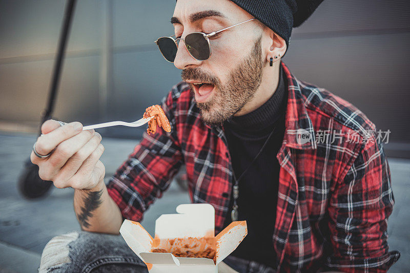 一个赶时髦的年轻人骑着电动滑板车坐在滑板车旁边休息，吃着美味的意大利面。他坐在人行道上，在商场的人行道上。