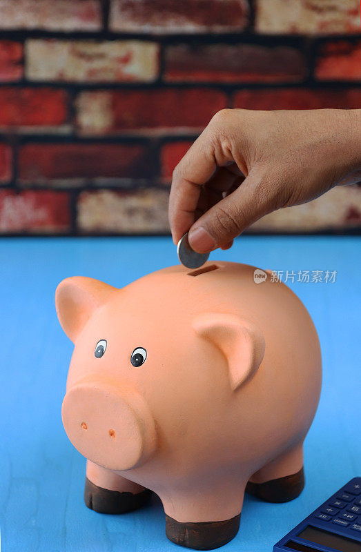 特写图像的不认识的人把一枚硬币放入槽上的小猪银行坐在计算器旁边，聚焦前景，家庭财务和储蓄的概念