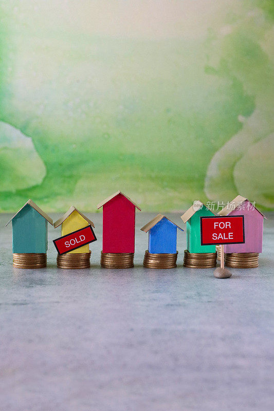 排着“待售”和“售出”标志的彩色纸板房子的图像，五颜六色的海滩小屋矗立在成堆的硬币上，重点放在前景，斑斑驳杂的绿色背景，家庭财务，房地产和假日储蓄概念