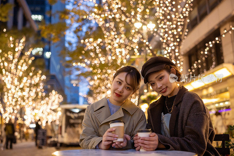 年轻的女性朋友在圣诞灯光下的街头咖啡馆里喝咖啡