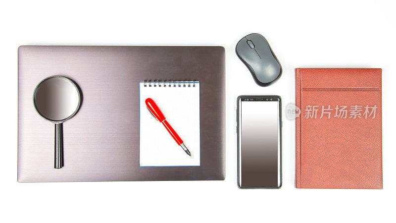 电脑无线鼠标，放大镜，智能手机，钢笔和记事本旁边的笔记本电脑在白色桌子上特写。办公室工作项目