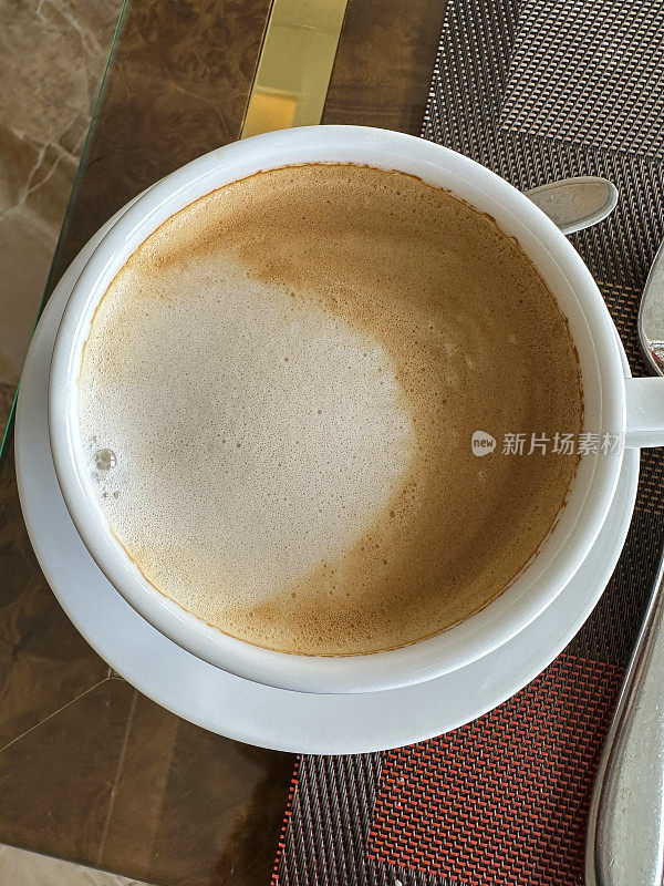 特写图像卡布奇诺咖啡在白色的杯子和碟子在餐厅的桌子上，早餐泡沫咖啡在餐垫，金属茶匙，抬高的观点